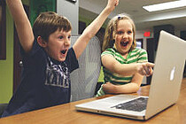 Foto zeigt zwei Kinder, ein Junge und ein Mädchen, die freudig lachend vor einem Notebook sitzend die Arme hochstrecken
